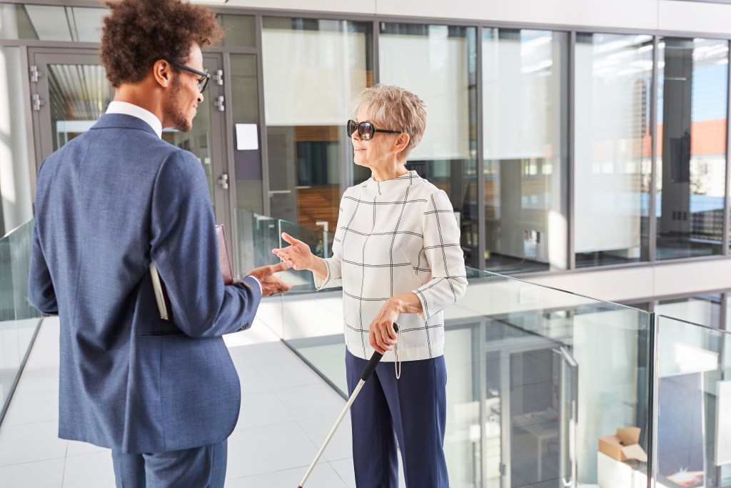 Two employees engaged in discussion in a brightly lit office space. One of the employees, a middle-aged woman with short, grey hair, uses a white cane.]
