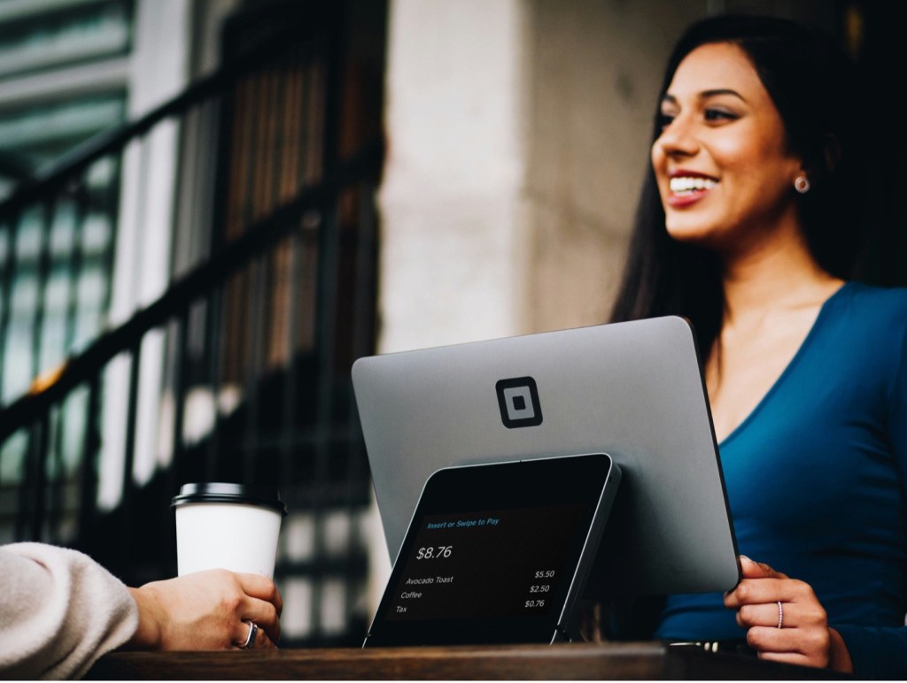 A smiling female cashier takes payment from a customer in a coffee shop.