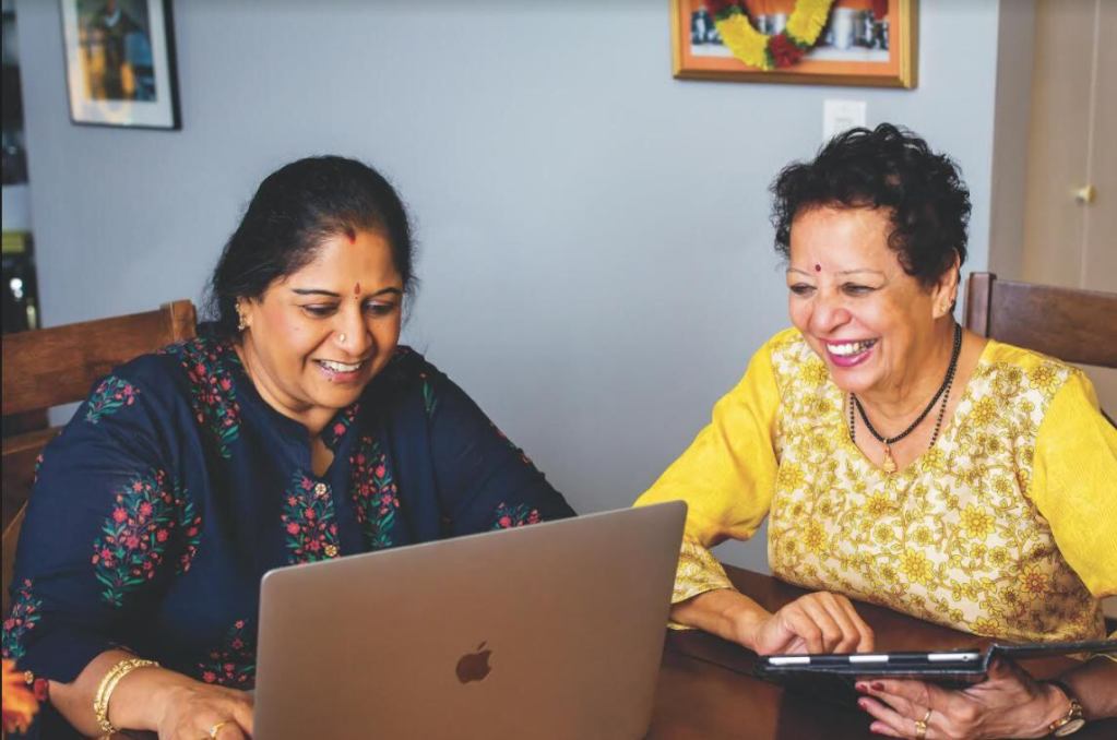 A smiling Prema and her friend Chamundi, type recipes together on a laptop. Photo courtesy of CBC News, and Baljit Singh.