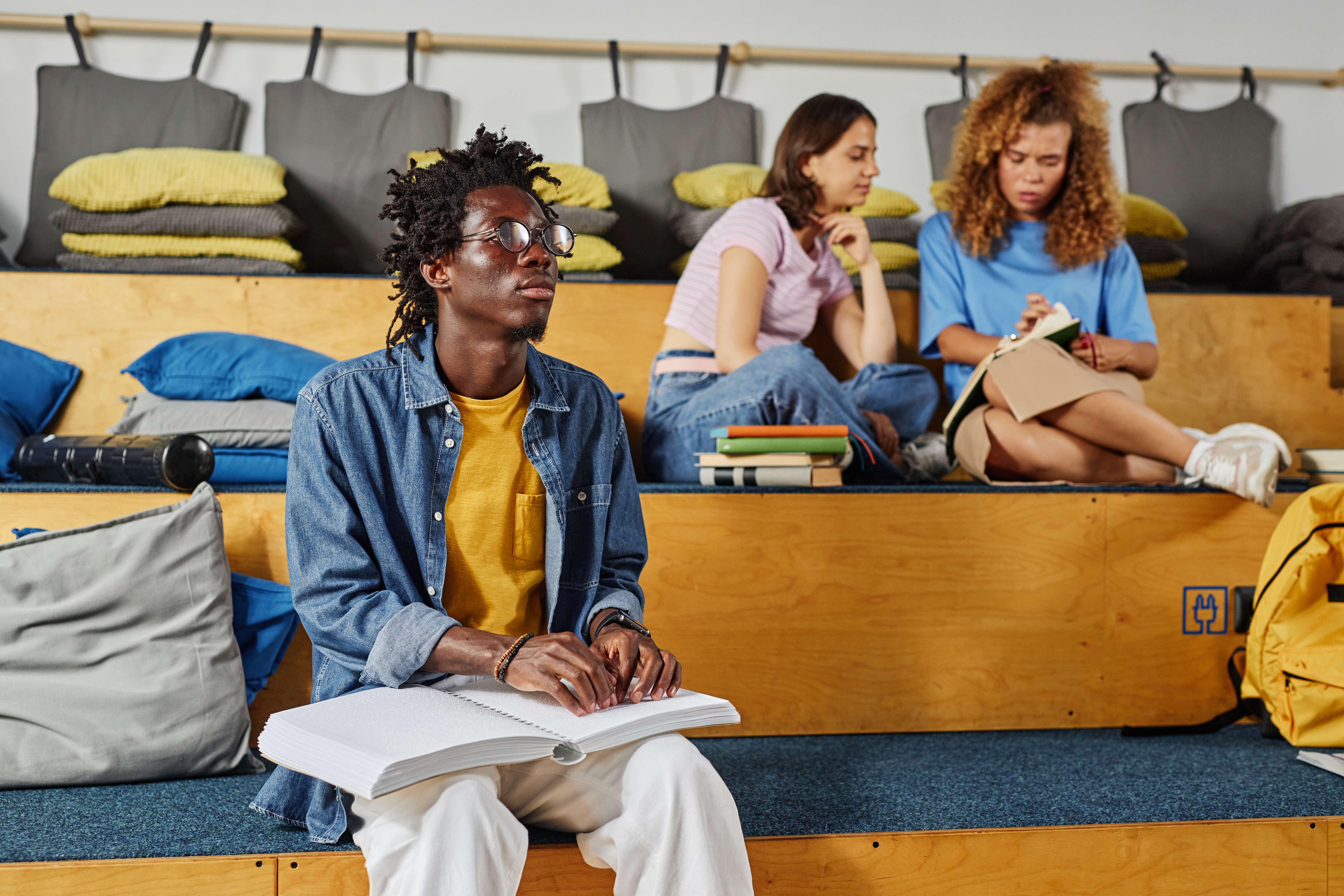 A young Black man reads a braille book while sitting in a common area of an educational institution. He is casually dressed in a denim shirt, a yellow t-shirt, and light khakis.