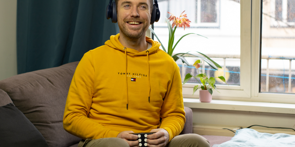 A young white man is enjoying music on his Hable One braille keyboard in his living room. He is wearing headphones and sitting on his couch.