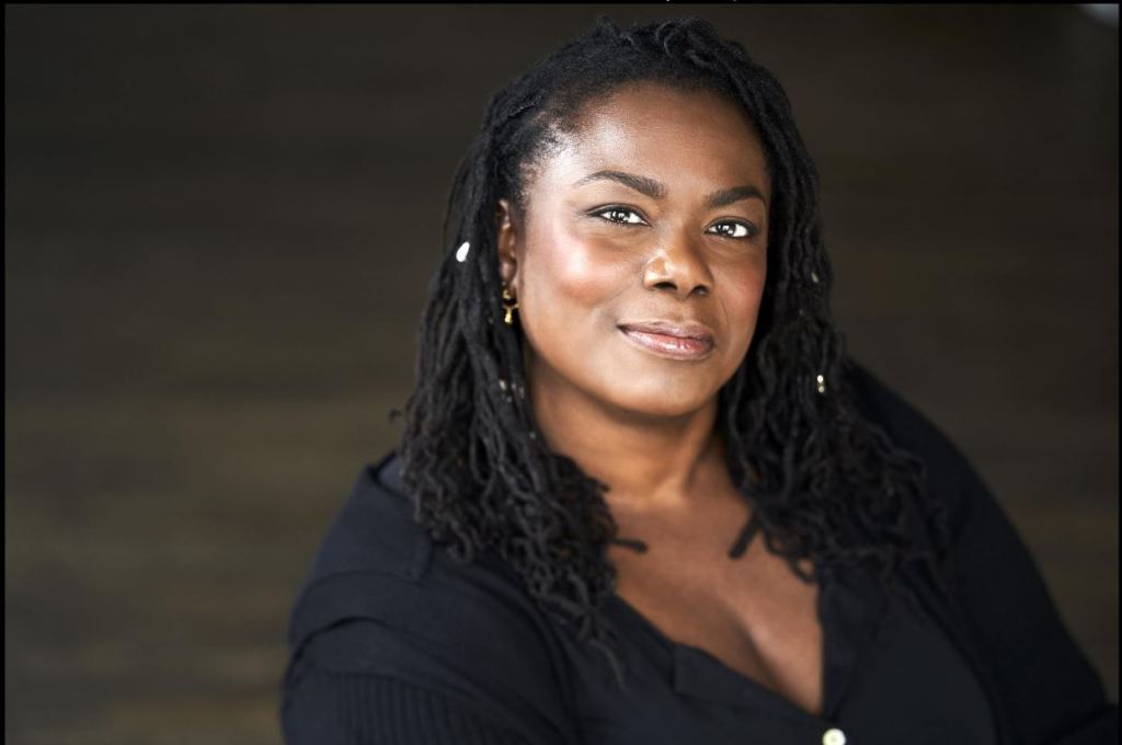 A professional headshot of Keda Edwards St. Pierre, wearing an elegant black button-down blouse against a dark background.